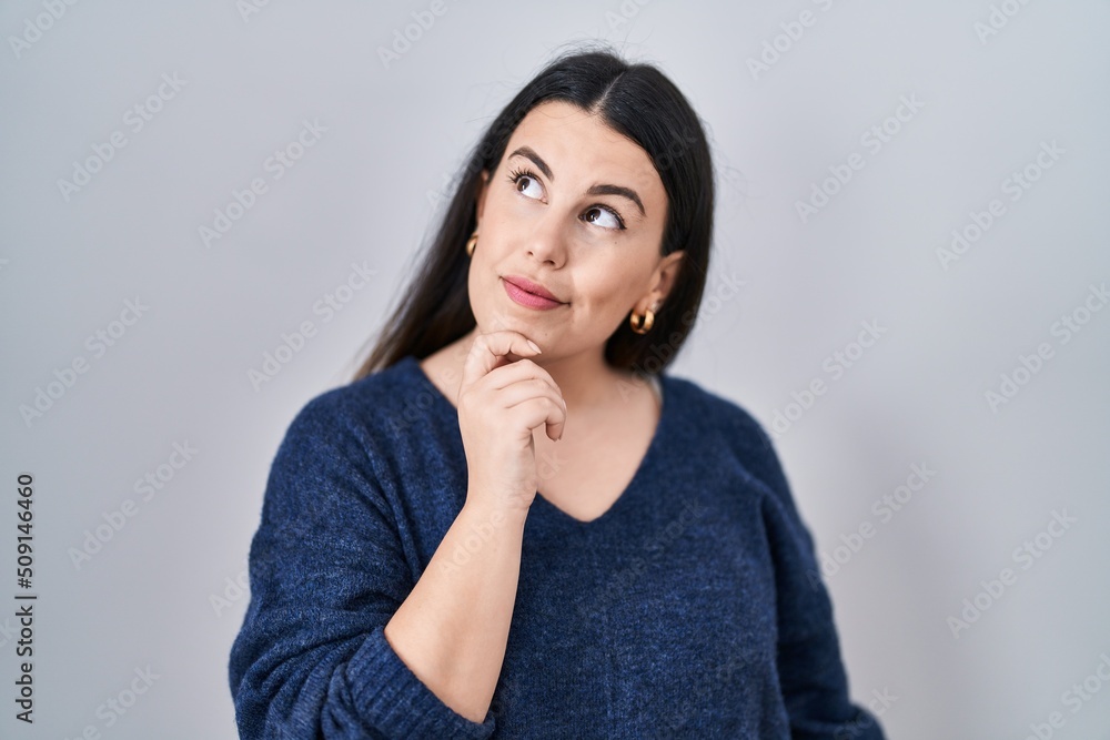 Young brunette woman standing over isolated background with hand on chin thinking about question, pensive expression. smiling with thoughtful face. doubt concept. Young brunette woman standing over isolated background with hand on chin thinking about question, pensive expression. smiling with thoughtful face. doubt concept.