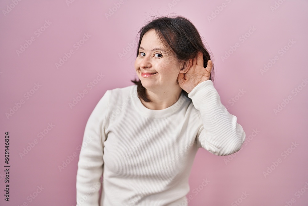 Woman with down syndrome standing over pink background smiling with hand over ear listening an hearing to rumor or gossip. deafness concept. Woman with down syndrome standing over pink background smiling with hand over ear listening an hearing to rumor or gossip. deafness concept.