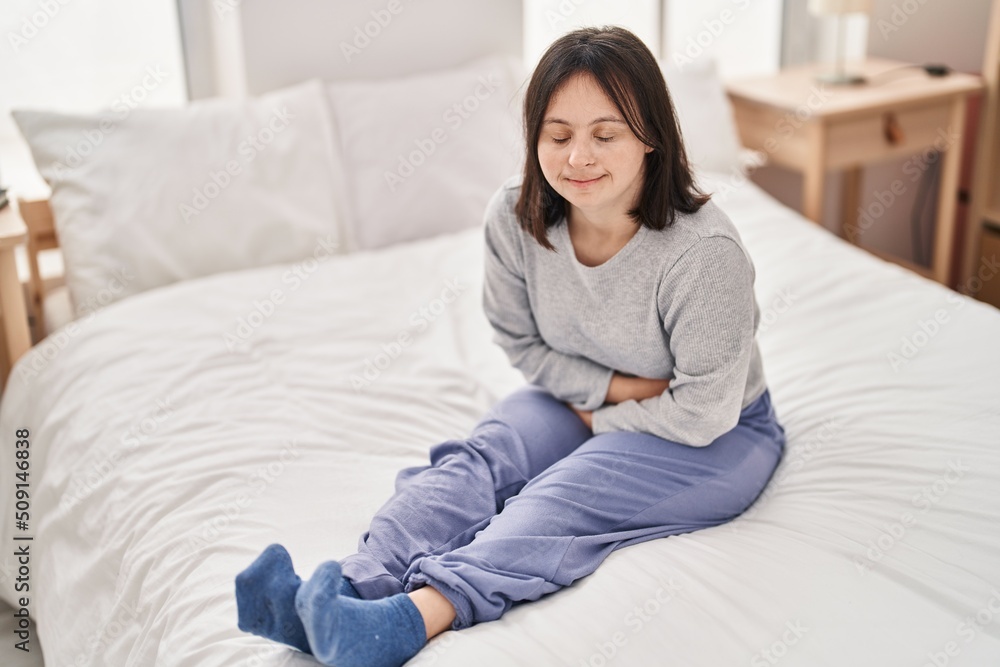 Young woman with down syndrome suffering for menstrual pain sitting on bed at bedroom Young woman with down syndrome suffering for menstrual pain sitting on bed at bedroom
