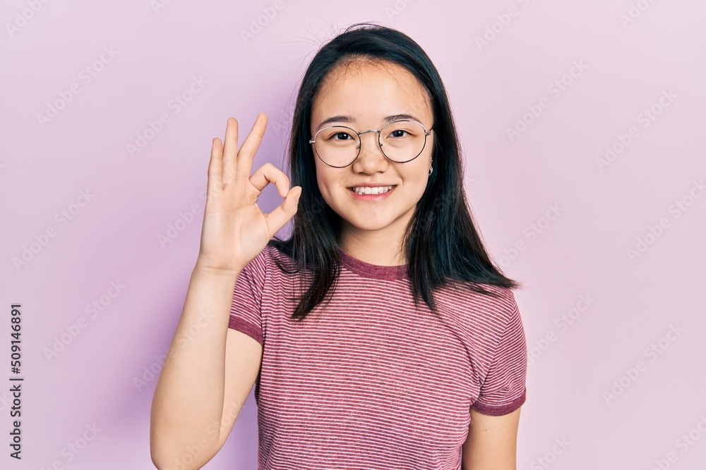 Young chinese girl wearing casual clothes and glasses smiling positive doing ok sign with hand and fingers. successful expression. Young chinese girl wearing casual clothes and glasses smiling positive doing ok sign with hand and fingers. successful expression.