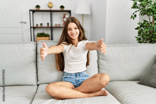 Young brunette teenager sitting on the sofa at home looking at the camera smiling with open arms for hug. cheerful expression embracing happiness.