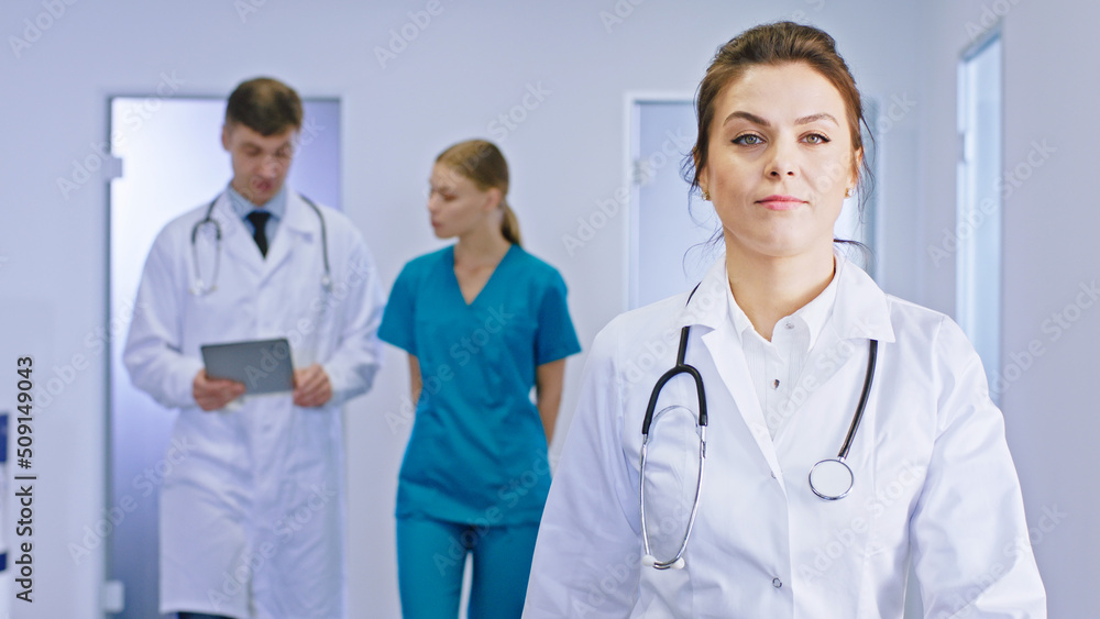 Mature woman doctor walking through the hospital corridor on the background other doctor man and his nurse lady discussing the future treatment for the patient Mature woman doctor walking through the hospital corridor on the background other doctor man and his nurse lady discussing the future treatment for the patient