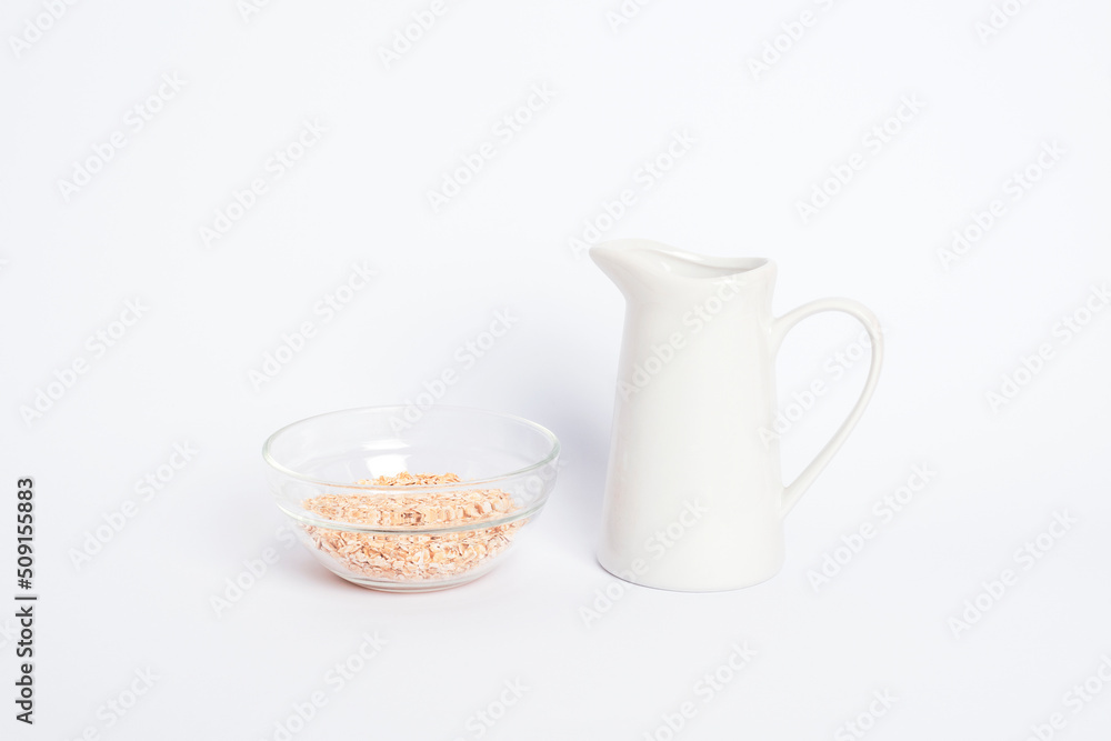 Jug of non dairy oat milk and oat flakes in glass bowl on white background. Healthy vegan food concept Jug of non dairy oat milk and oat flakes in glass bowl on white background. Healthy vegan food concept