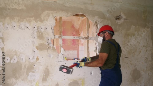 Contractor wrecks wall with sledgehammer making hole for rearrangement. Man doing manual dismantling and demolition works with big hammer hits for apartment renovation. Construction worker in uniform. photo