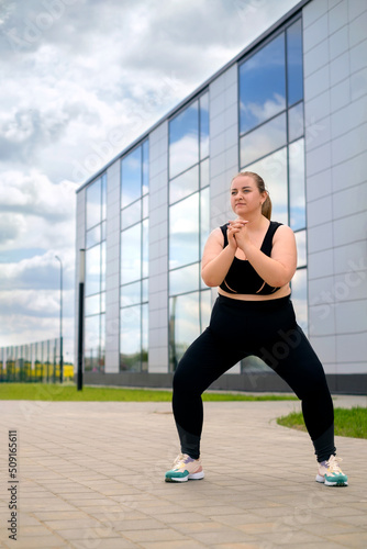 Young plus-size woman does sports on street in city on sidewalk. Fitness outdoors. Fatty chubby plump female in black tracksuit trains doing squats in fresh air at modern building. World Obesity Day