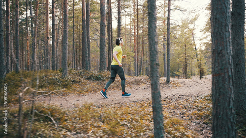 Man in bright yellow t-shirt runs through pine forest. Young man in white headphones runs on forest path for cardio training slow motion