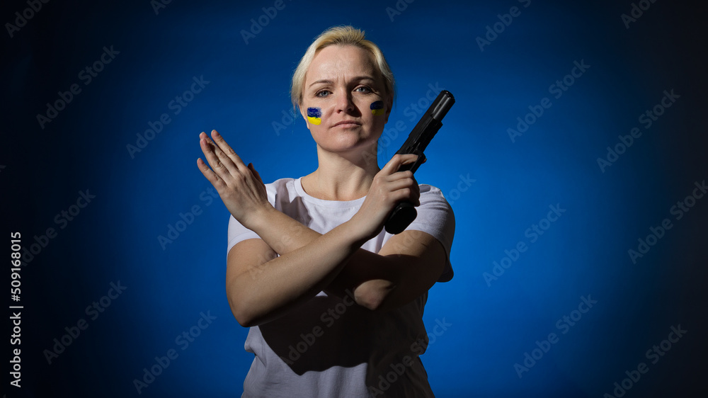 Fototapeta premium a woman with a gun and a Ukrainian flag on her face, crossed her arms in a stop sign on a dark background