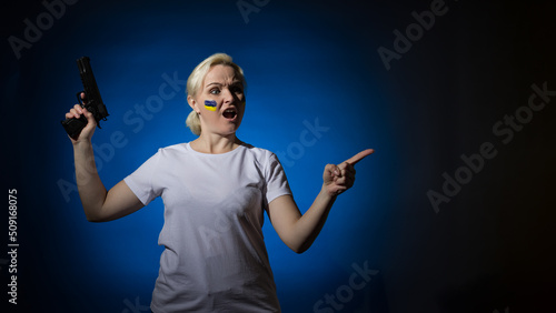 An aggressive woman with a gun and with a blue and yellow Ukrainian flag on her cheek points her finger to the side. Dark background. Place for text.