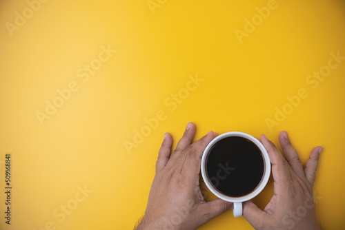 Hand of a man holding a white cup with hot black coffee on yellow background with copy space. Morning drinks coffee. top view espresso coffee break.