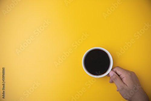 Hand of a man holding a white cup with hot black coffee on yellow background with copy space. Morning drinks coffee. top view espresso coffee break.