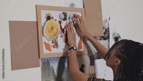 Close up view of African American woman attaching paper element to canvas in flame while making creative collage on wall in art studio photo