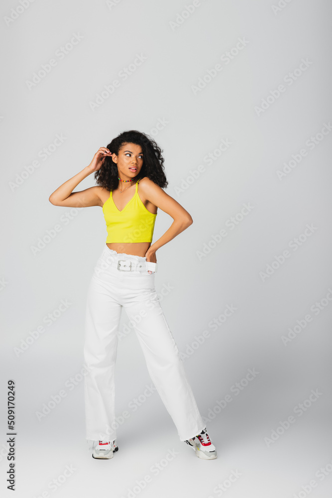 full length of african american woman in trendy outfit and stylish sneakers posing with hand on hip isolated on grey. full length of african american woman in trendy outfit and stylish sneakers posing with hand on hip isolated on grey.