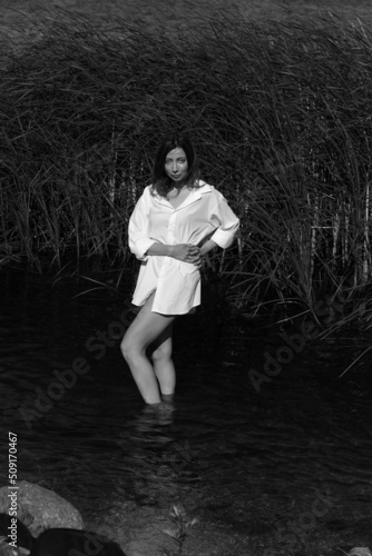 Beautiful woman in shirt standing in water near reeds in black and white