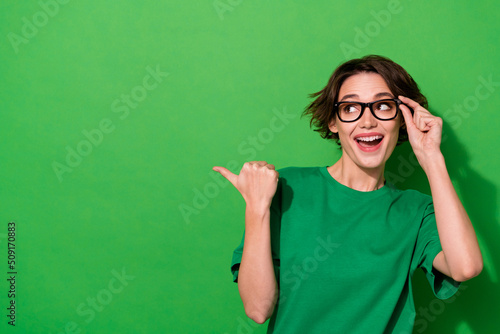 Photo of adorable funny woman wear casual t-shirt eyeglasses pointing thumb looking empty space isolated green color background