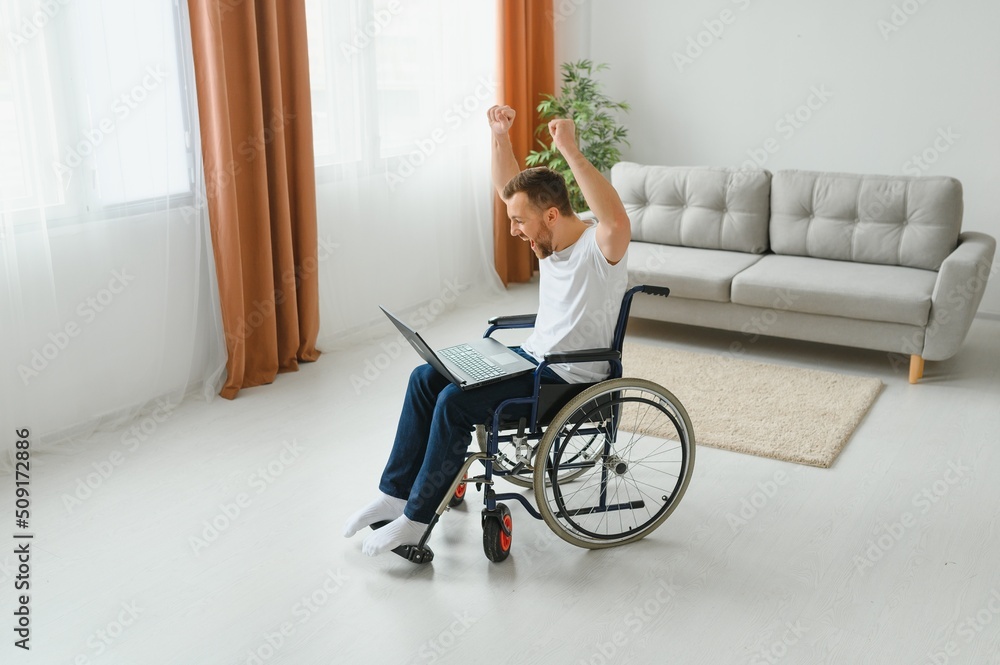 Portrait of smiling disabled male sitting in wheelchair and working on laptop from home. Young worker with special needs. Freelancer and people with disabilities concept Portrait of smiling disabled male sitting in wheelchair and working on laptop from home. Young worker with special needs. Freelancer and people with disabilities concept
