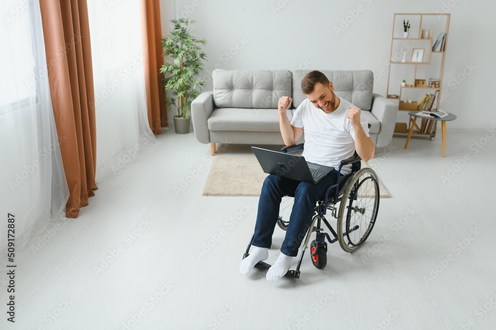Portrait of smiling disabled male sitting in wheelchair and working on laptop from home. Young worker with special needs. Freelancer and people with disabilities concept Portrait of smiling disabled male sitting in wheelchair and working on laptop from home. Young worker with special needs. Freelancer and people with disabilities concept