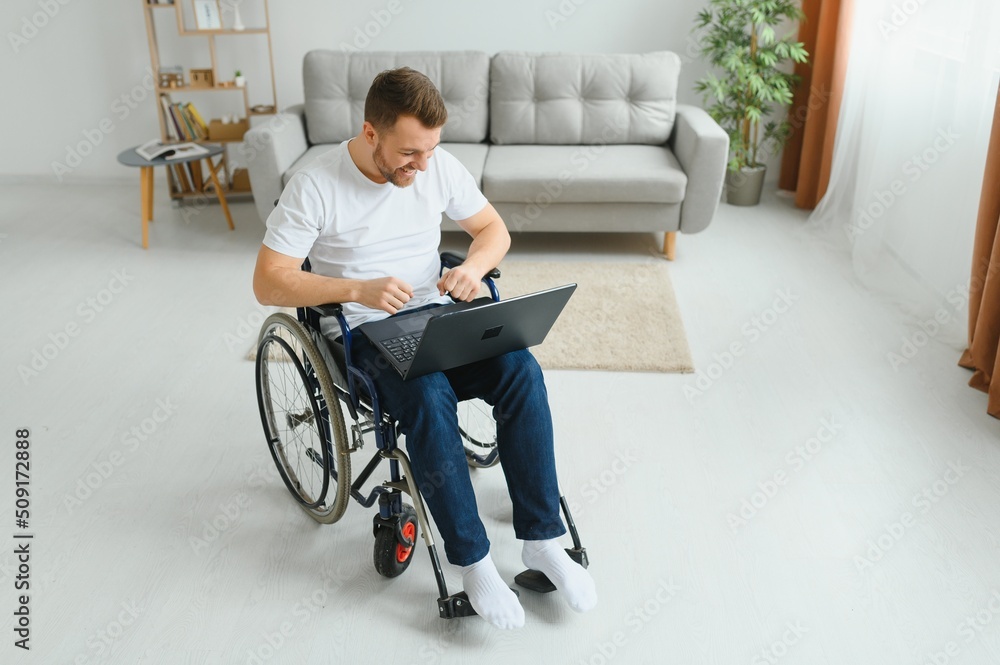Portrait of smiling disabled male sitting in wheelchair and working on laptop from home. Young worker with special needs. Freelancer and people with disabilities concept Portrait of smiling disabled male sitting in wheelchair and working on laptop from home. Young worker with special needs. Freelancer and people with disabilities concept