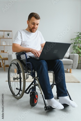 Portrait of smiling disabled male sitting in wheelchair and working on laptop from home. Young worker with special needs. Freelancer and people with disabilities concept