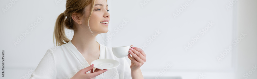Smiling woman holding coffee cup and saucer at home, banner.