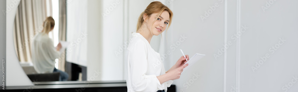 Blonde woman smiling at camera and writing on notebook at home, banner. Blonde woman smiling at camera and writing on notebook at home, banner.