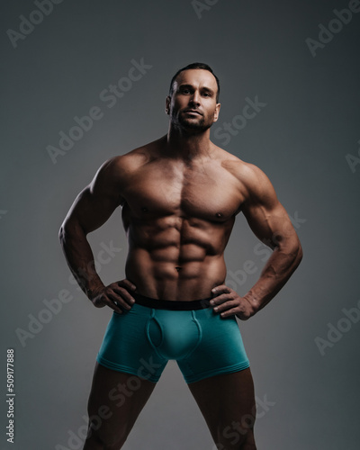 Muscular sexy guy in turquoise shorts standing with hands on his belt in studio