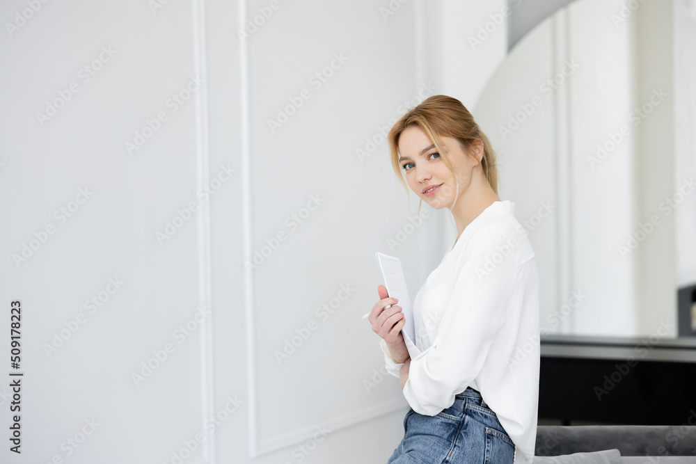 Happy young woman holding notebook and looking at camera in living room. Happy young woman holding notebook and looking at camera in living room.