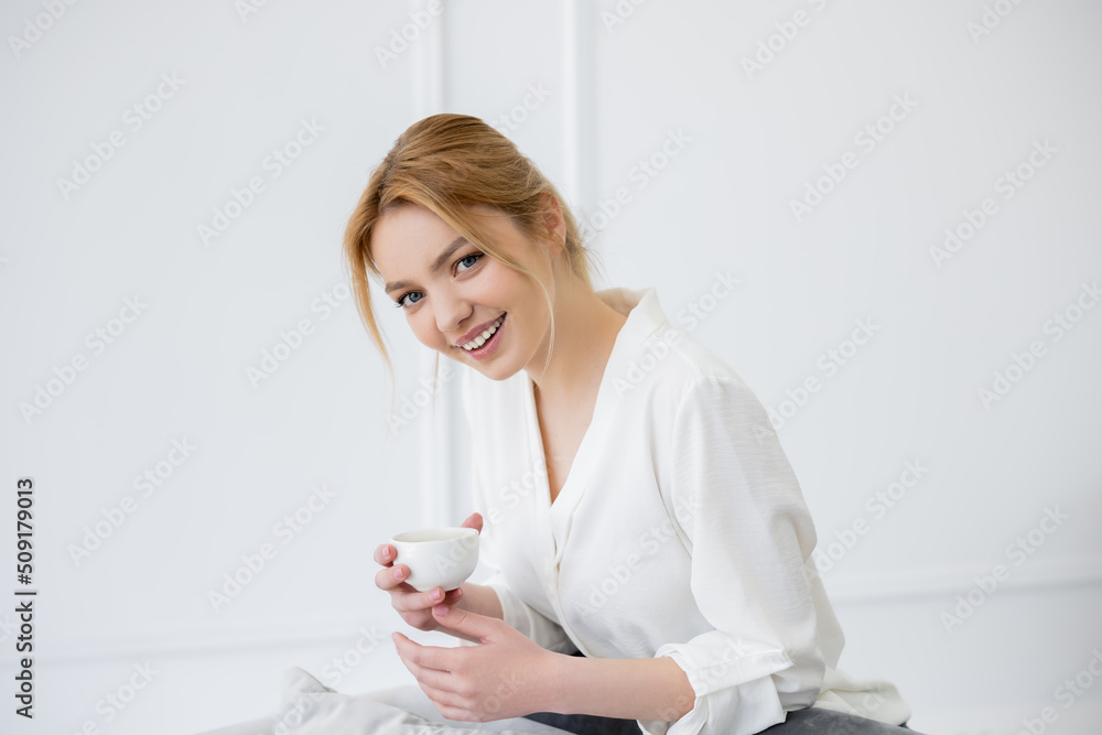 Happy young woman in blouse holding coffee cup and looking at camera at home. Happy young woman in blouse holding coffee cup and looking at camera at home.