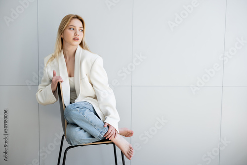 Fotografie Barefoot woman in jeans and jacket sitting on chair at home.