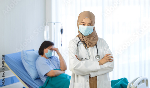 Young muslim female doctor arms crossed stand in hospital with elderly patient lie down on bed on the background.
