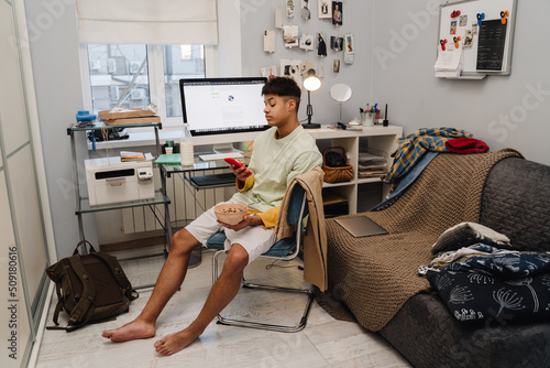 Brunette teenage boy using cellphone and eating breakfast in bedroom