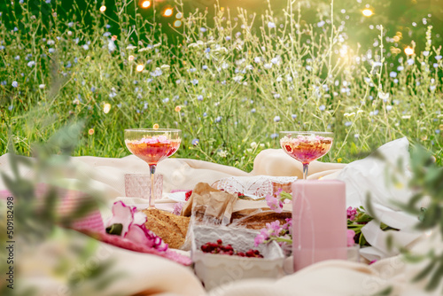 Aesthetic summer sunny lunch picnic with baguette, flowers, cocktail and berries at pale beige blanket at grass outdoor background with sunbeam. Front view. photo