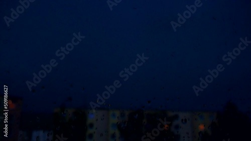 Thunderstorm at night with lots of lightning in the sky over the city behind the window. Focus purposefully on the window with raindrops so that the city and storm in the background were blured