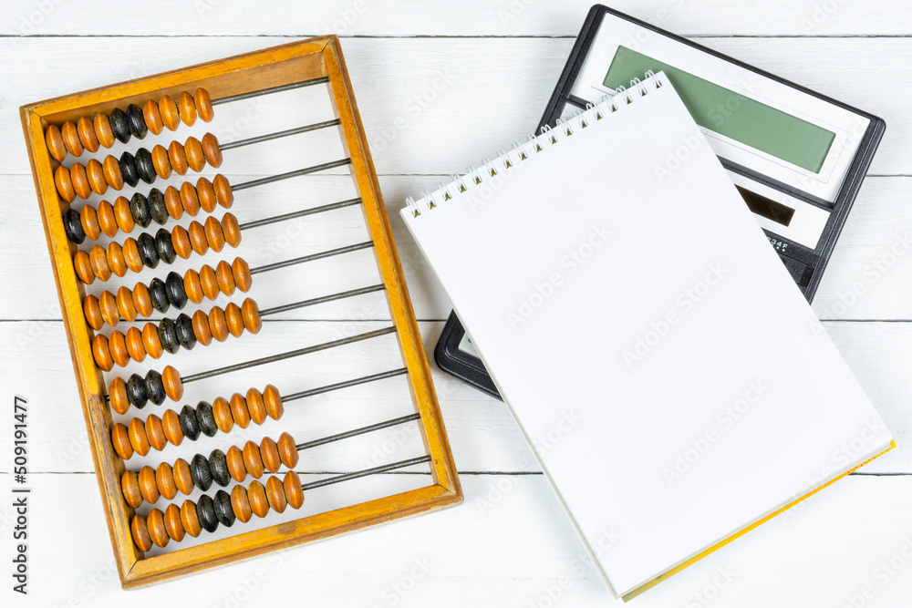Old antique wooden abacus, a modern black calculator and a notebook with a blank page for writing on a white wooden background. Space for text. Old antique wooden abacus, a modern black calculator and a notebook with a blank page for writing on a white wooden background. Space for text.
