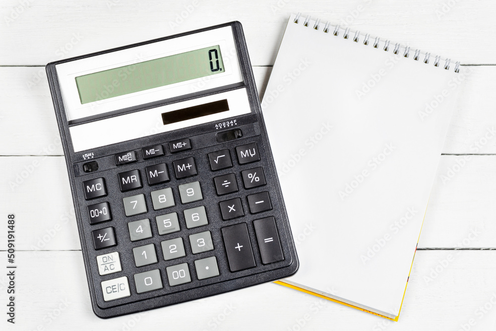 A modern black calculator and notepad (notebook) with a blank page for writing on a white wooden background. A modern black calculator and notepad (notebook) with a blank page for writing on a white wooden background.