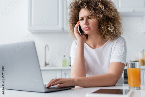 serious and frowning woman using laptop and talking on cellphone in kitchen.