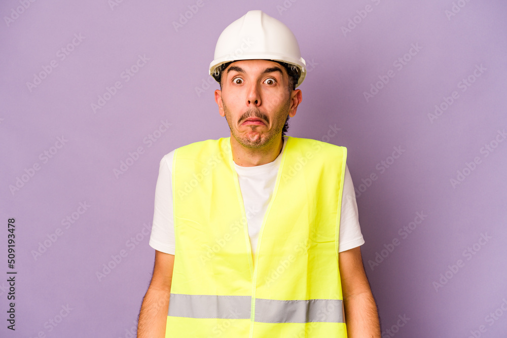 Young hispanic worker man isolated on purple background shrugs shoulders and open eyes confused.