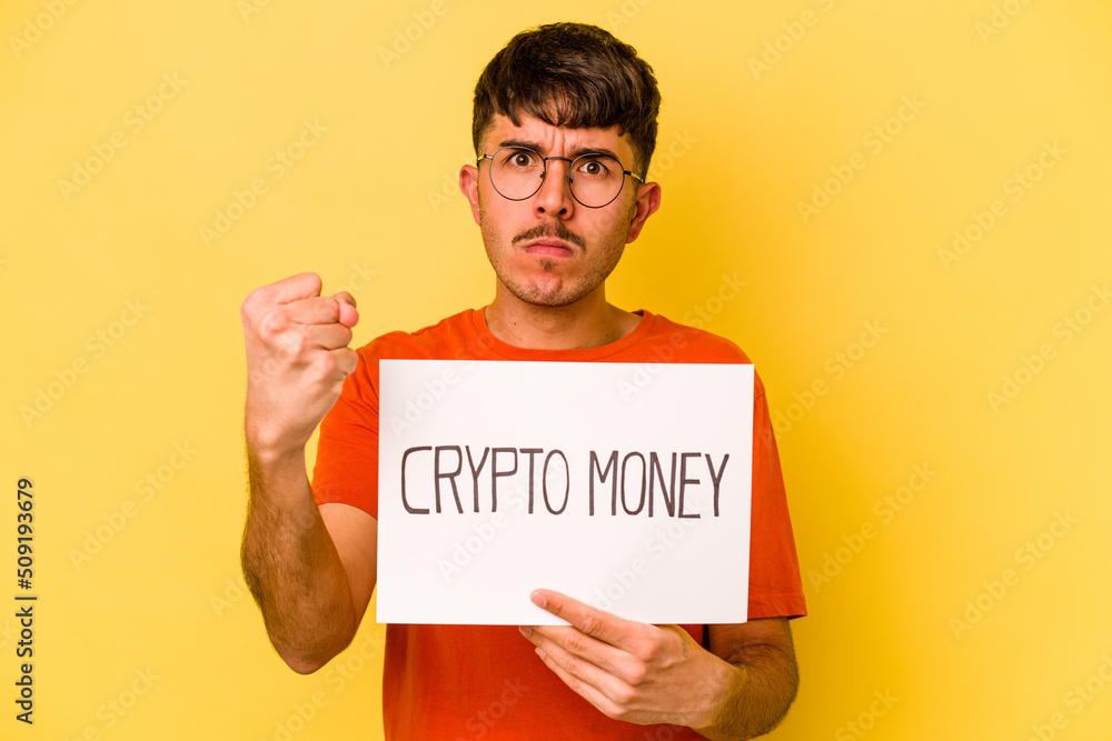 Young hispanic man holding nft placard isolated on yellow background showing fist to camera, aggressive facial expression. Young hispanic man holding nft placard isolated on yellow background showing fist to camera, aggressive facial expression.