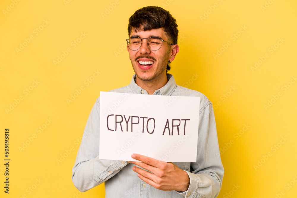 Young hispanic man holding crypto art placard isolated on yellow background laughs out loudly keeping hand on chest. Young hispanic man holding crypto art placard isolated on yellow background laughs out loudly keeping hand on chest.