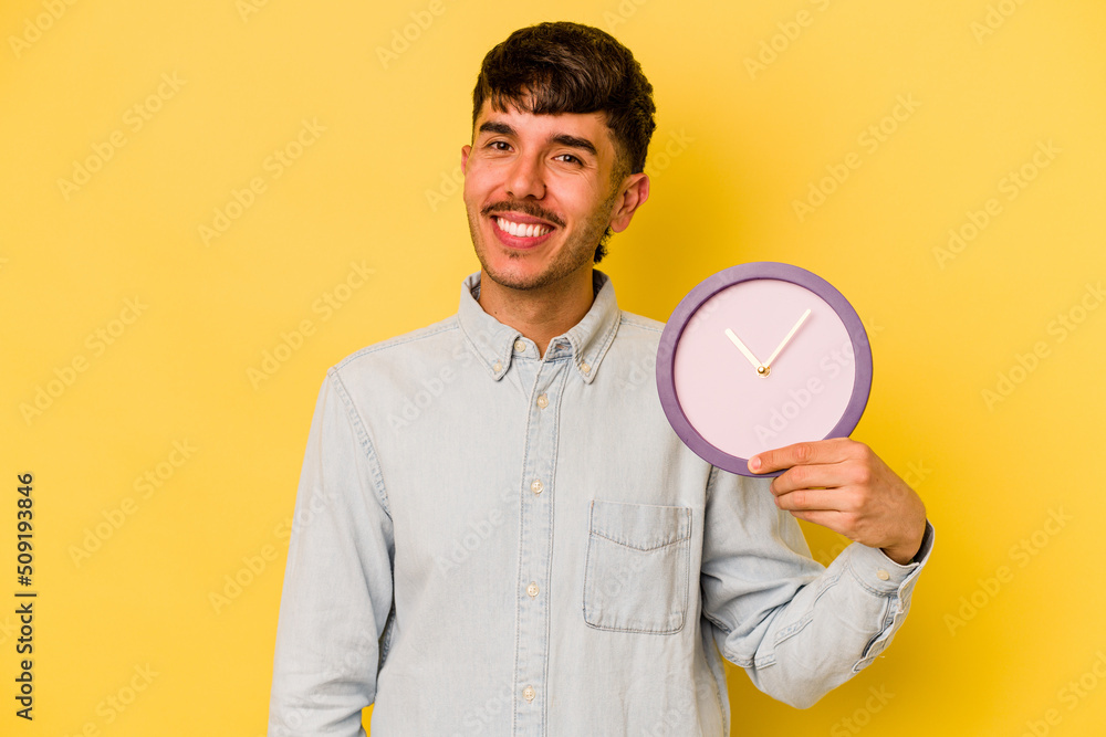 Young hispanic man holding a clock isolated on yellow background happy, smiling and cheerful. Young hispanic man holding a clock isolated on yellow background happy, smiling and cheerful.