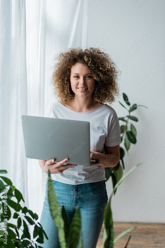 happy and curly woman standing with laptop at home and looking at camera. happy and curly woman standing with laptop at home and looking at camera.