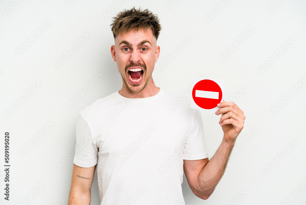 Young caucasian man holding a forbidden sign isolated on white background screaming very angry and aggressive. Young caucasian man holding a forbidden sign isolated on white background screaming very angry and aggressive.