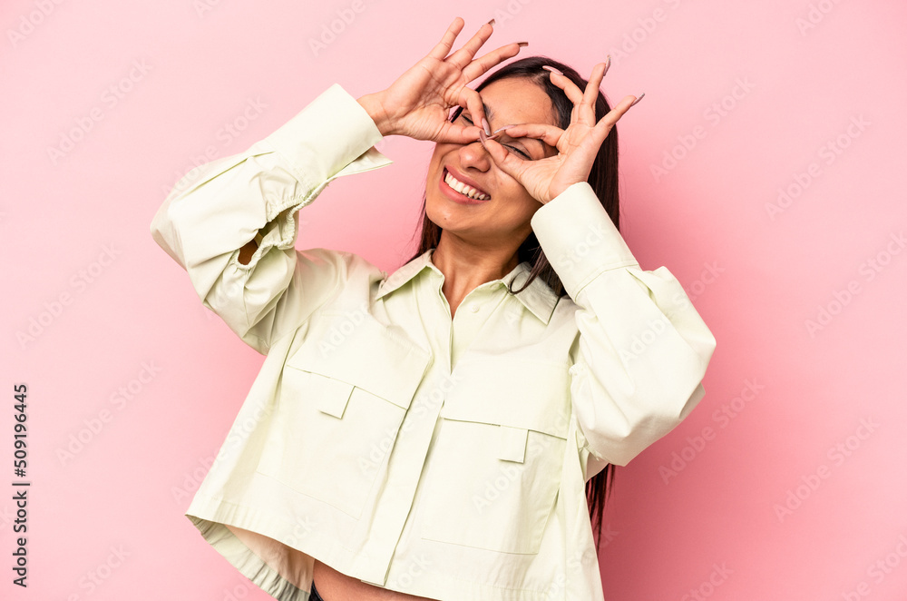Young hispanic woman isolated on pink background showing okay sign over eyes