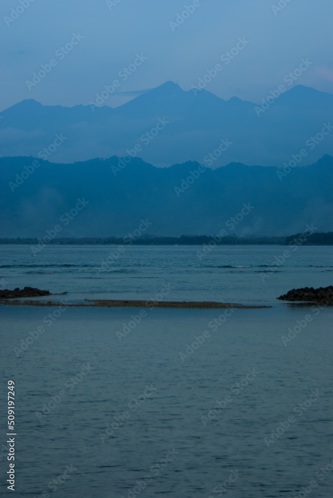 Blue evening on sea with mountains in fog on horizon in calm cloudy sunset, vertical. Overcast landscape on indonesian island, travel on asia. Blue evening on sea with mountains in fog on horizon in calm cloudy sunset, vertical. Overcast landscape on indonesian island, travel on asia.