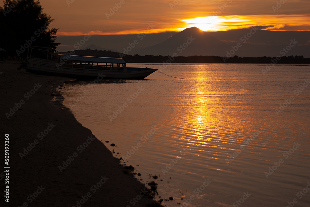 Scenic golden sea sunset with dark volcano in distance island with colorful orange heaven and sunbeams reflection in water, silhouette boat on black shore. Majestic indonesian landscape, trip in asia.