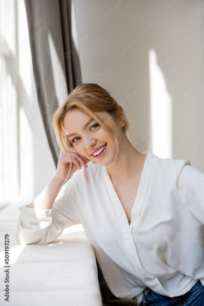 Positive young woman looking at camera near windowsill. Positive young woman looking at camera near windowsill.