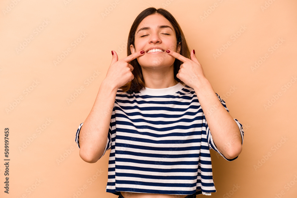 Young hispanic woman isolated on beige background smiles, pointing fingers at mouth. Young hispanic woman isolated on beige background smiles, pointing fingers at mouth.