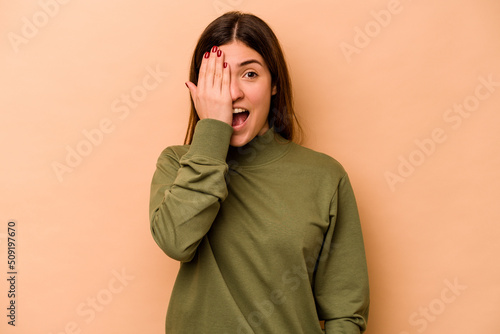 Young hispanic woman isolated on beige background having fun covering half of face with palm.