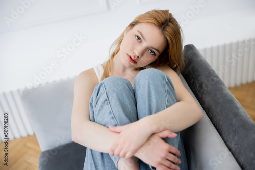 Portrait of young woman in blurred jeans looking at camera while sitting on couch.