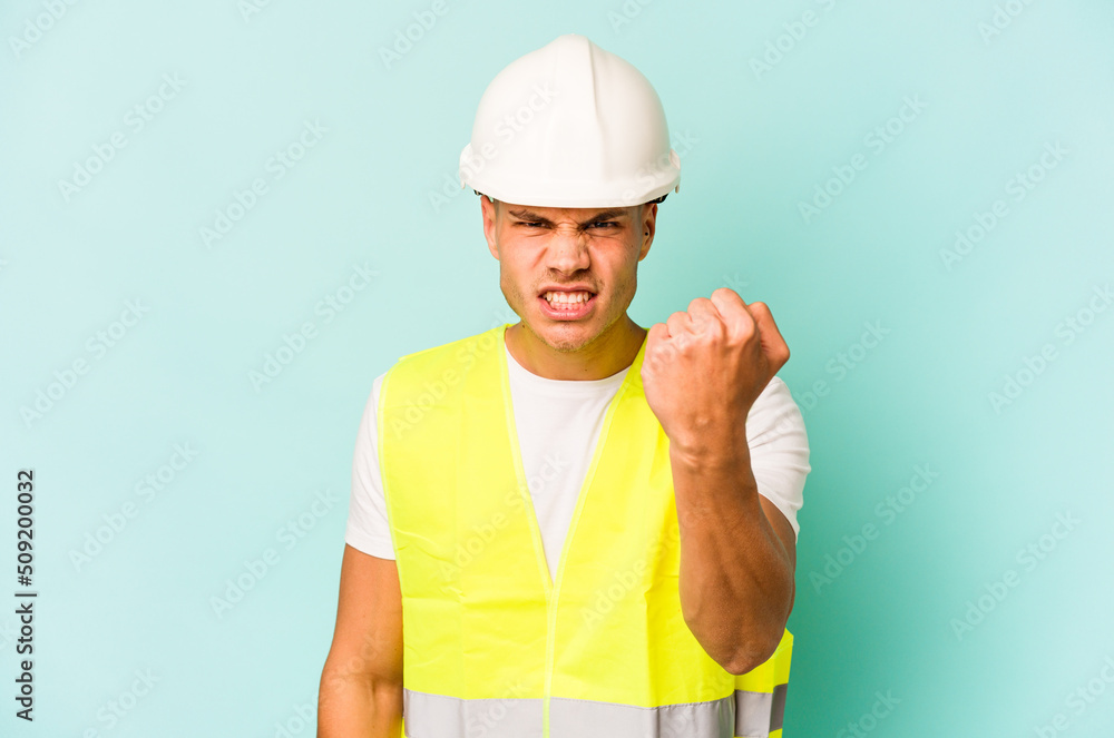 Young laborer caucasian man isolated on blue background showing fist to camera, aggressive facial expression. Young laborer caucasian man isolated on blue background showing fist to camera, aggressive facial expression.