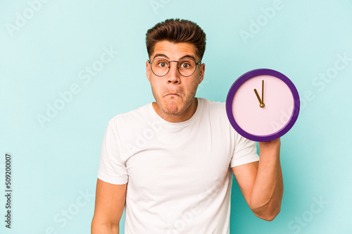 Картината върху платно Young caucasian man holding a clock isolated on blue background shrugs shoulders and open eyes confused